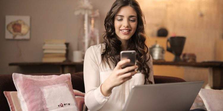 Woman Sitting on Sofa While Looking at Phone With Laptop on Lap